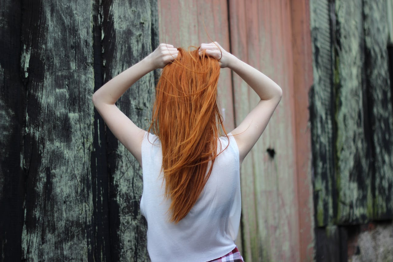 services-01 A red-haired woman poses with her back to the camera, outdoors against a rustic wooden backdrop.