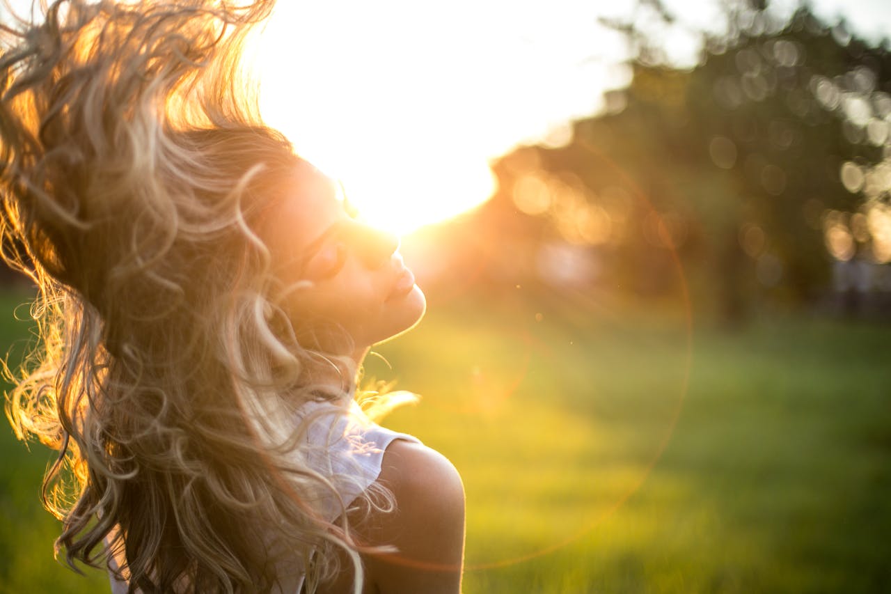 who-we-are A young woman tosses her long, wavy blond hair in the sunlight during golden hour outdoors.